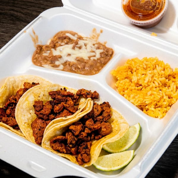 Three tacos with meat, rice, refried beans, lime wedges, and two sauces in a takeout container.