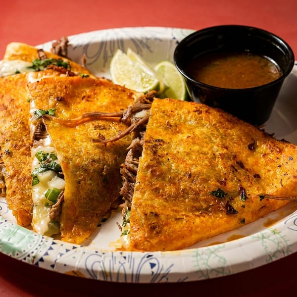 Three golden-brown birria tacos with melted cheese, shredded beef, lime wedges, and a side of consommé.