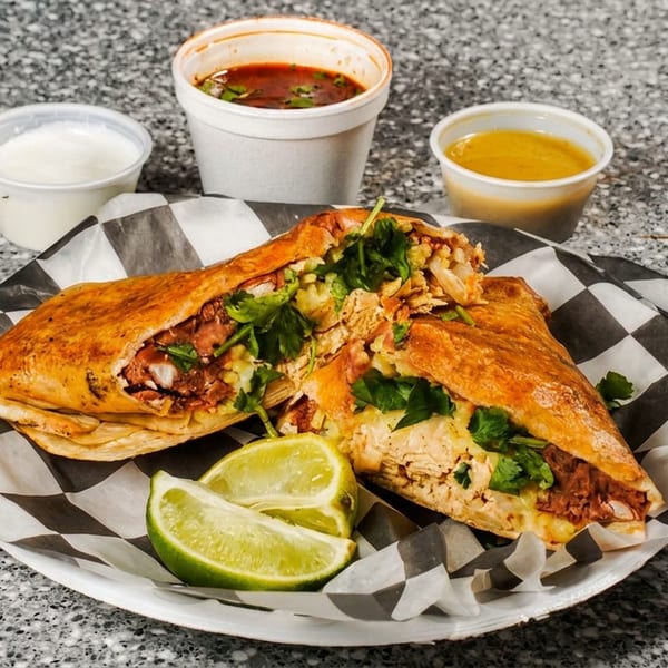 Two halves of a crispy burrito filled with meat and cilantro, with limes, sauces, and soup.