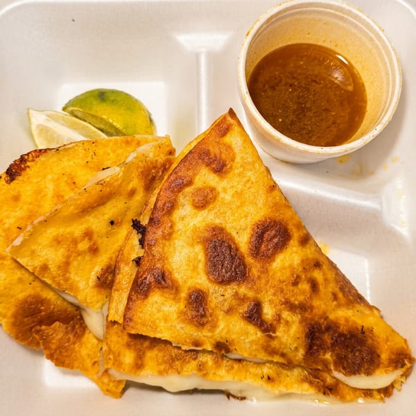 Three cheesy quesadilla slices with lime wedges and a side of dipping sauce in a to-go container.
